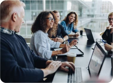 Stock photo of a group of people varying in age, race, and gender collaborating around a table using their paper and electronic notebooks