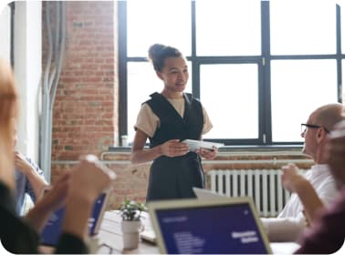 Stock photo of a group of people varying in age, race, and gender collaborating around a table, where a woman is standing and talking to an older man